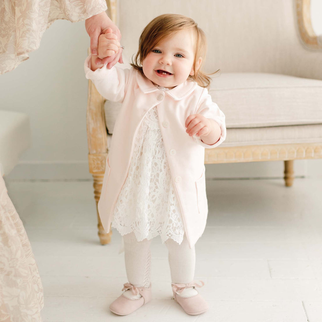 Baby girl wearing the Lola Trench Coat in soft pink, layered over the Lola Lace Romper. The cotton French terry coat features a petite collar, button closures, and front pockets, while she holds an adult's hand and smiles.