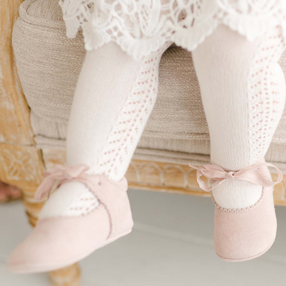 Close-up of a baby girl wearing the Lola Suede Tie Mary Janes, made from soft rosy pink suede with scalloped edge detailing and finished with a delicate cotton ribbon tie.