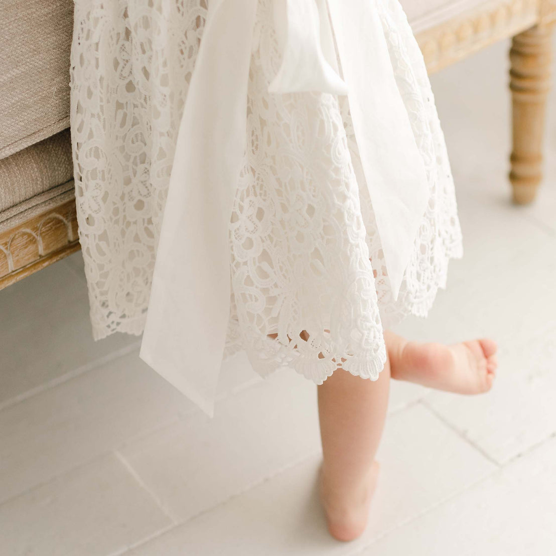 A baby standing up and wearing the Lola Dress with attention to the details of the skirt and sash on the back. The baby appears to be standing against a upholstered chair.