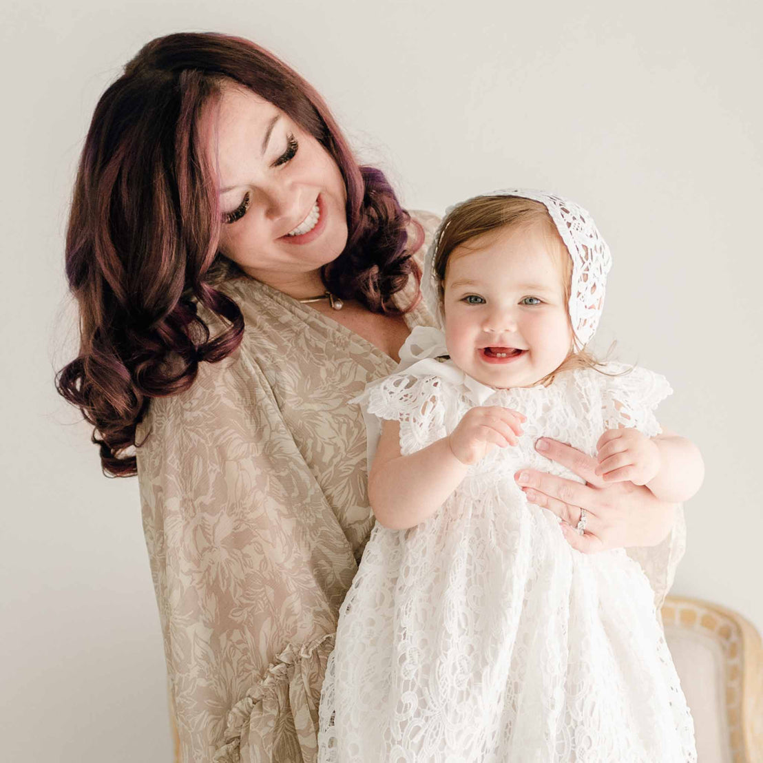 Mother holding her smiling baby girl dressed in the Lola Christening Gown and Bonnet. The daughter looks to the side with her a slight smile as her mother looks down upon her.