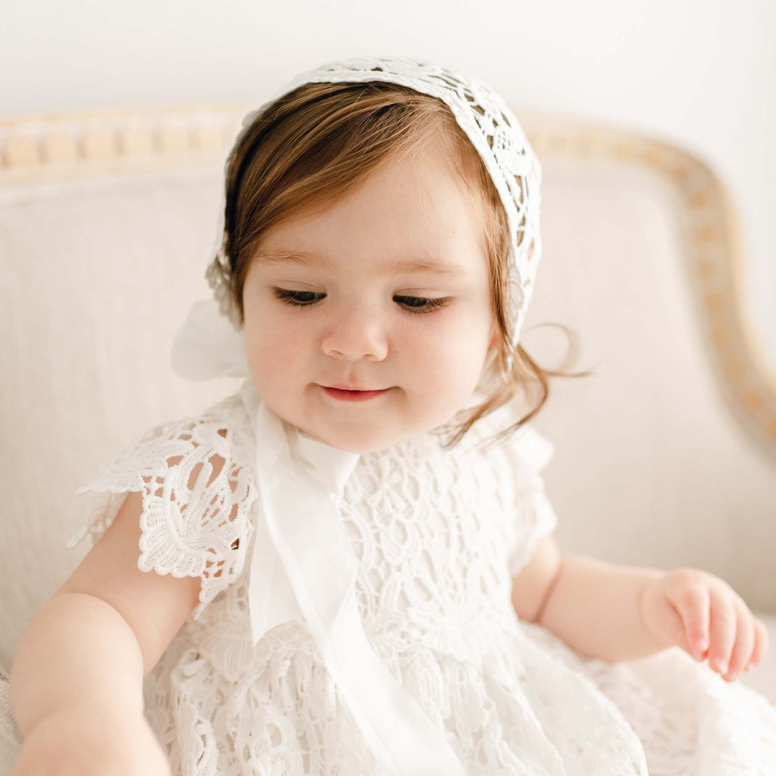 Close up of a baby girl with red hair wearing the Lola Christening Gown and Bonnet. The baby girl is looking down with a slight smirk on her face. The baptismal gown and bonnet features an all-over lace overlay.