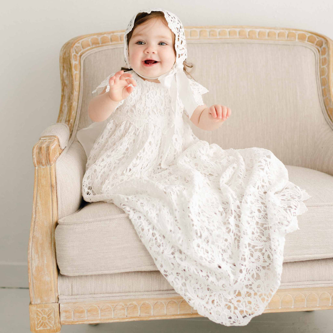 A baby girl sitting on a linen chair and wearing the Lola Christening Gown and Bonnet, a baptismal gown an all-over lace overlay. She appears holding up her arms and gazing to the side with a smile.