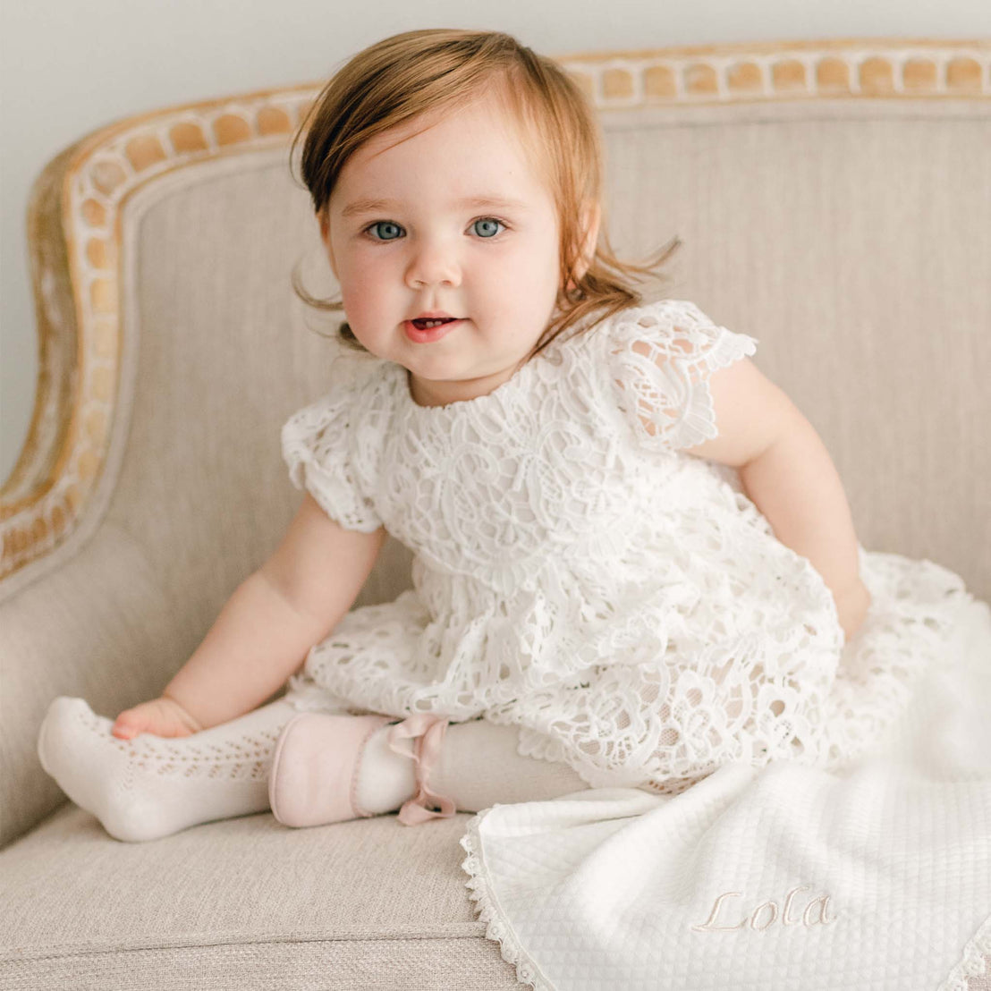 A baby girl sitting on an upholstered linen chair with a Lola Personalized Blanket draped to the side of the seat. On one foot she is wearing one Lola Pink Suede Tie Mary Jane shoe.