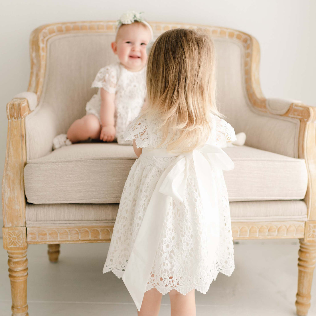 Two girls, one sitting on an upholstered linen chair and the other standing, appear to be looking at each other. The toddler girl in front is wearing the Lola Dress with details showing the back sash. The other baby girl is wearing the Lola Lace Bubble Romper and sitting on a upholstered linen chair and smiling.