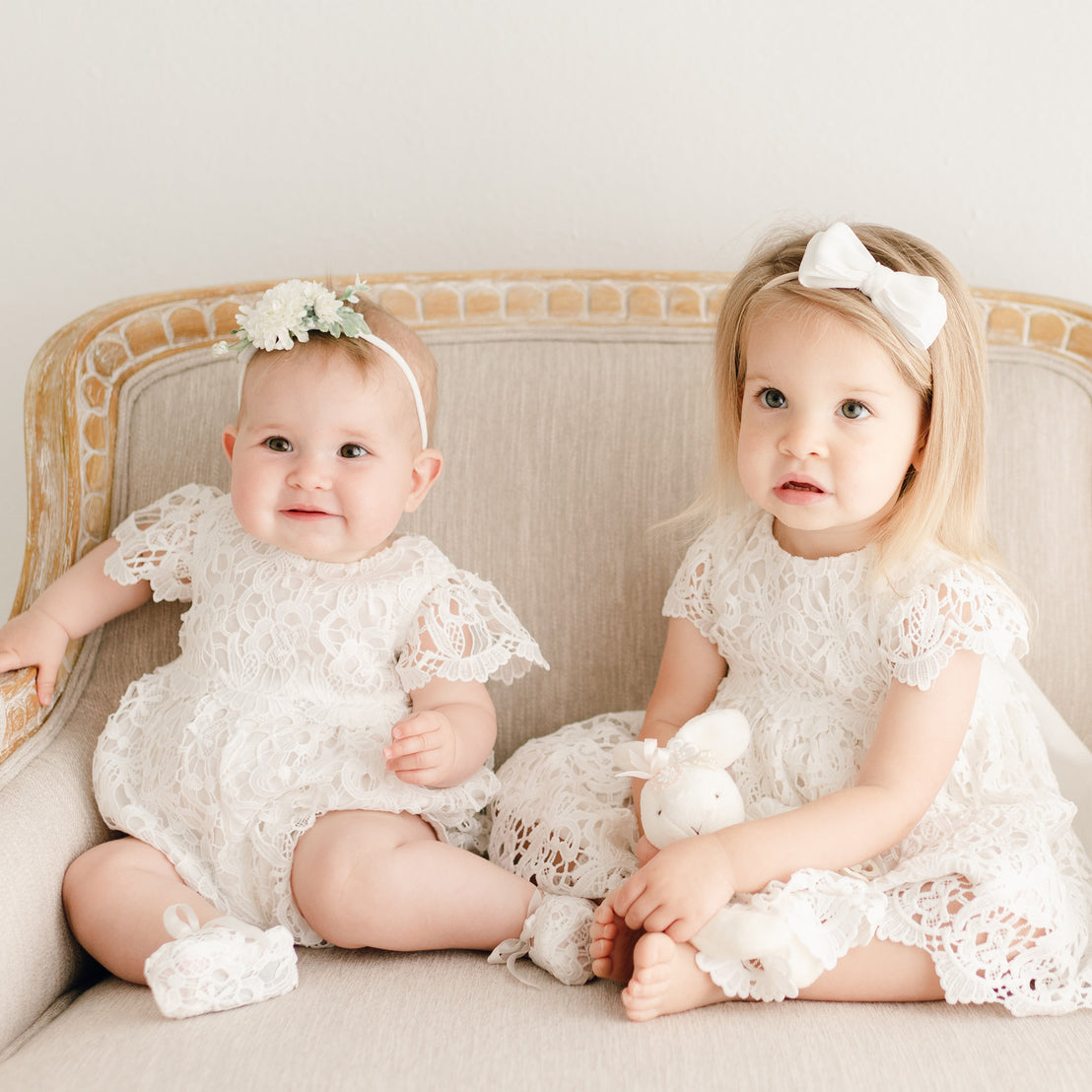 Two baby girls sitting on a beige chair, both wearing the Lola Lace Bubble Romper. One is styled with the Lola Flower Headband, featuring cream flowers and greenery, while the other is wearing a white bow headband.