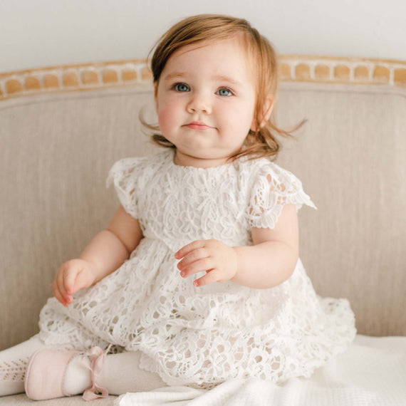 A baby girl wearing the Lola fancy baby dress sits on an upholstered linen chair with a slight grin on her face.