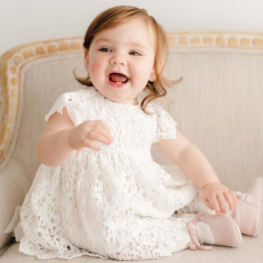 A baby girl sitting on a linen chair and smiling with a big open mouth grin. She is wearing the boutique all-over lace Lola Dress and pink suede Mary Janes.