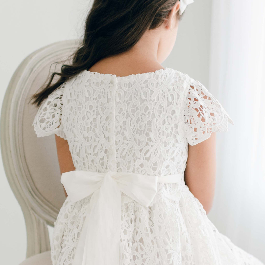 Back view of a girl wearing the Lola Girls Lace Dress, featuring intricate lace detail and a large bow tied at the waist. The light ivory lace and cap sleeves create a timeless, elegant look for special occasions.