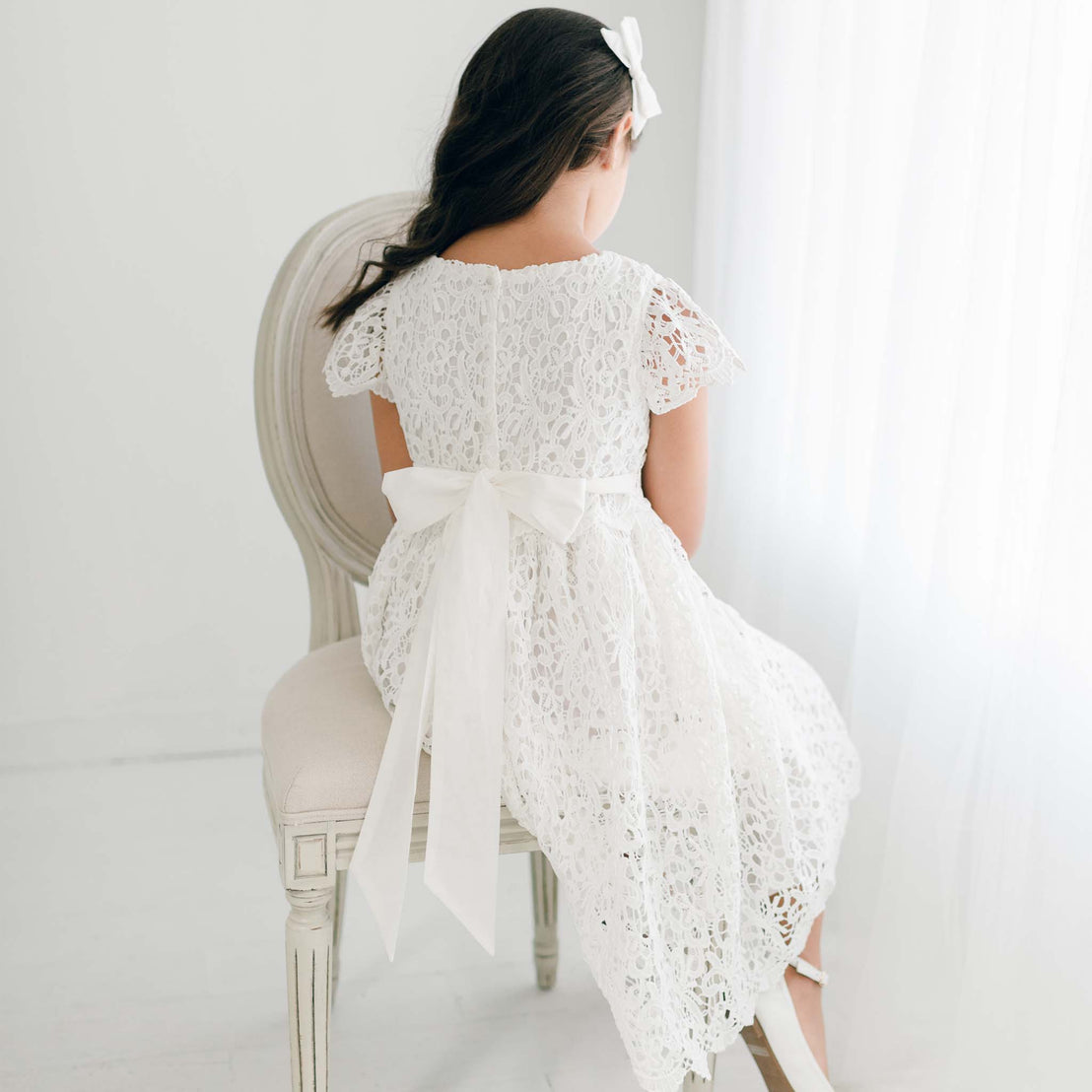 Back view of a young girl wearing the Lola Girls Lace Dress in light ivory, featuring a delicate all-over lace design, cap sleeves, and a large bow tied at the waist. The dress is completed with a button-back closure and a matching white bow headband.