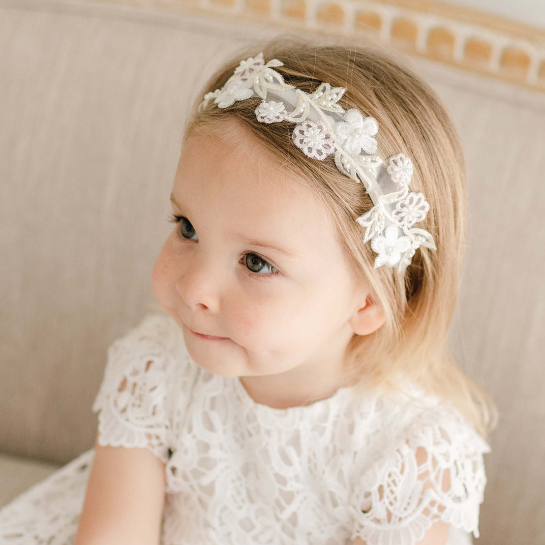 Baby girl wearing the Lola Beaded Flower Headband, featuring delicate floral appliqué in light ivory with beaded accents, paired with a matching lace dress.