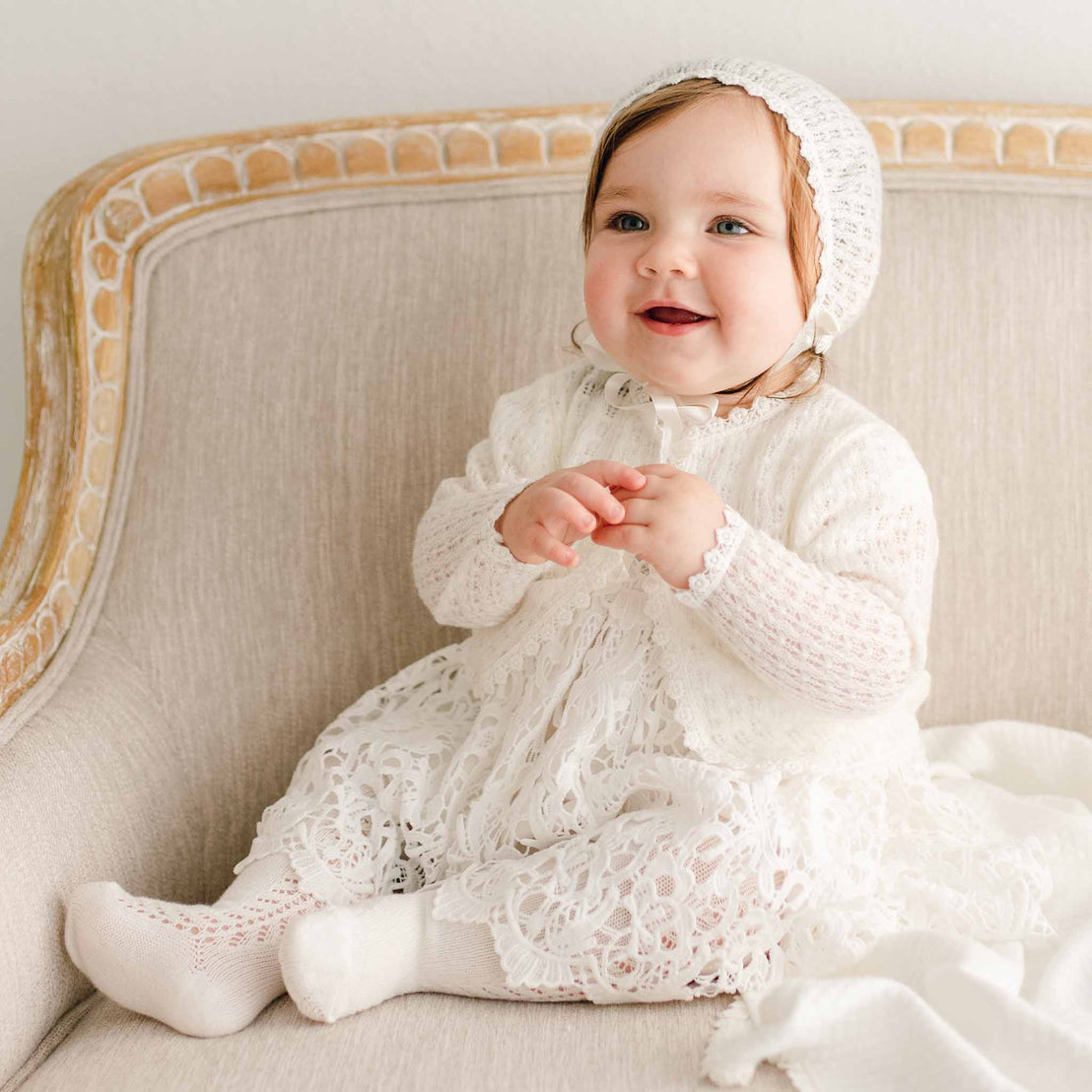 Baby girl sitting on a beige chair, wearing the Lola Knit Bonnet with light ivory knit fabric, delicate lace trim, and silk ribbon ties, paired with the matching lace christening gown and knit sweater.