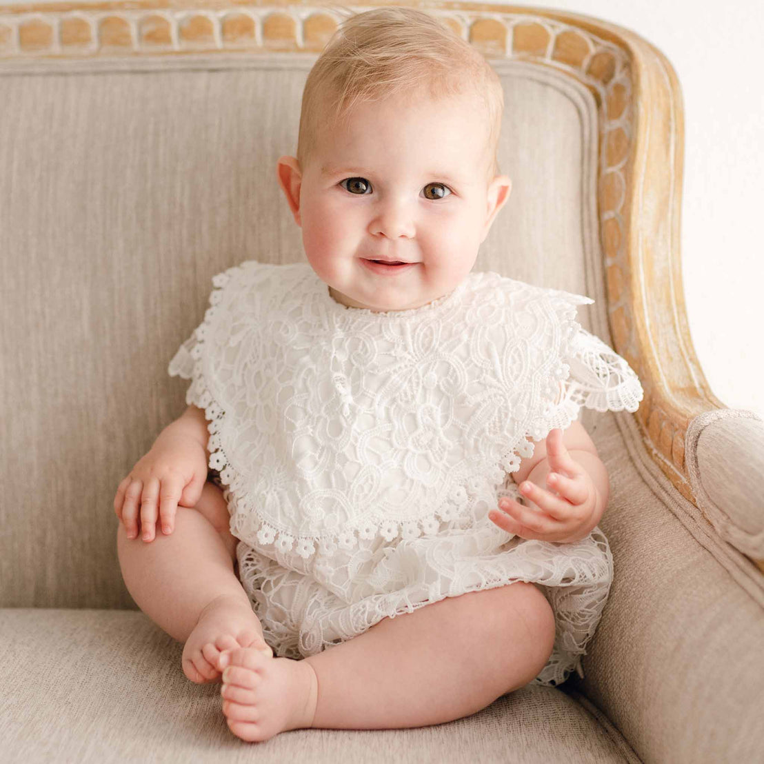 Baby girl sitting on a beige chair, wearing the Lola Bib featuring delicate light ivory lace and cotton lace trim, paired with a matching lace romper.