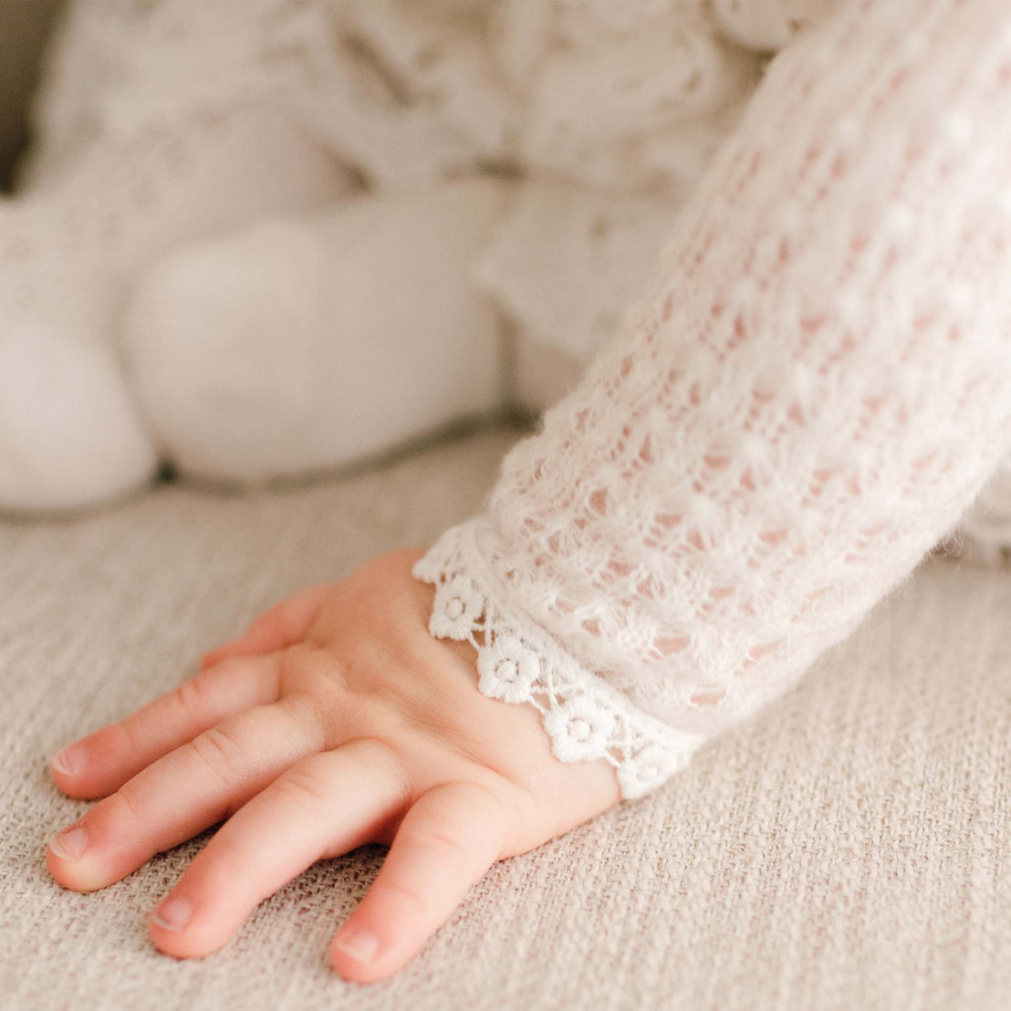 Close-up of a baby’s hand resting on fabric, wearing the Lola Knit Sweater with intricate light ivory knit fabric and lace-trimmed cuffs.