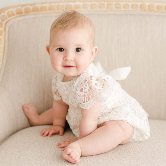 Baby girl wearing the Lola Lace Bubble Romper, featuring delicate light ivory floral lace, seated on a linen-upholstered chair.