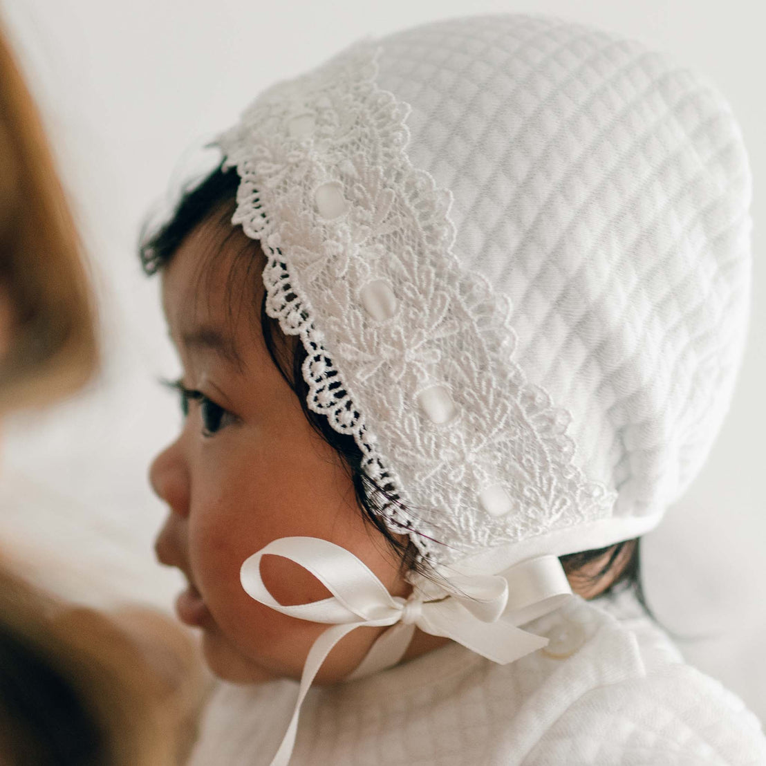 A baby wearing a Madeline Quilted Newborn Bonnet adorned with lace and decorative buttons looks to the side. The quilted cotton bonnet is tied under the chin with silk ribbon ties. The baby has dark hair and is dressed in a matching white outfit.