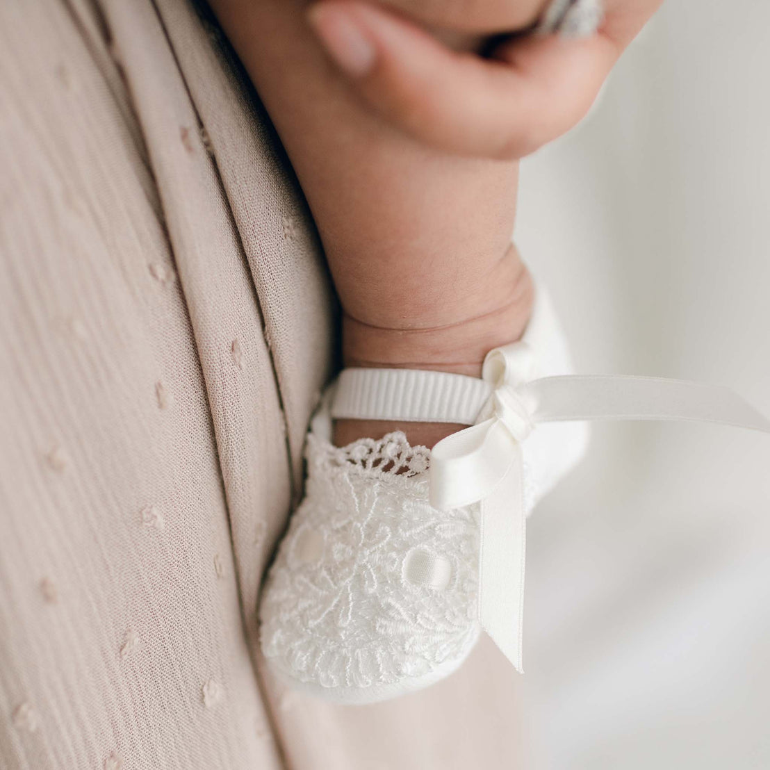 Close-up of a baby's foot wearing Madeline Quilted Booties with a small ribbon bow. The baby is held gently by an adult, with a soft, textured beige blanket visible in the background. The charming baby girl booties showcase delicate ivory lace, adding to their timeless appeal.