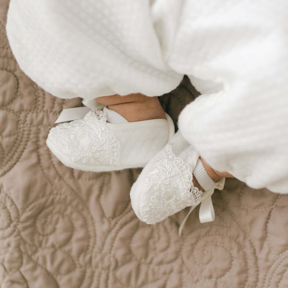 A close-up photo of a baby wearing white lace-trimmed shoes and white pants. The Madeline Quilted Booties rest against a quilted, beige blanket.