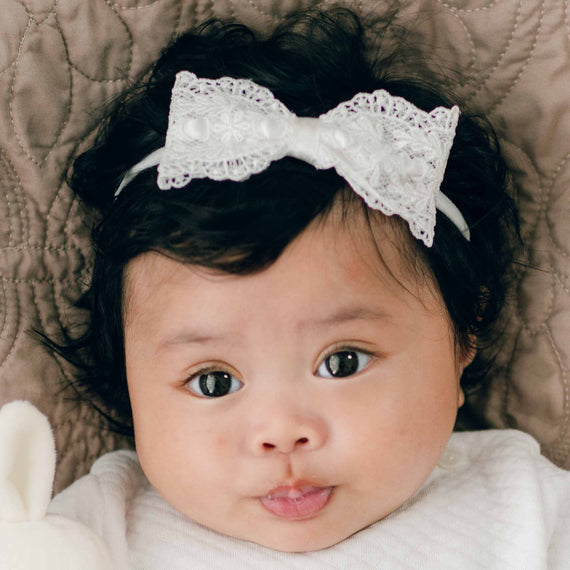 A close-up of a baby with dark curly hair, wearing the Madeline Lace Bow Headband. The baby is lying on a textured quilted surface and is looking up with wide eyes and a slightly open mouth. The baby, dressed in a white outfit, exudes charm and innocence.