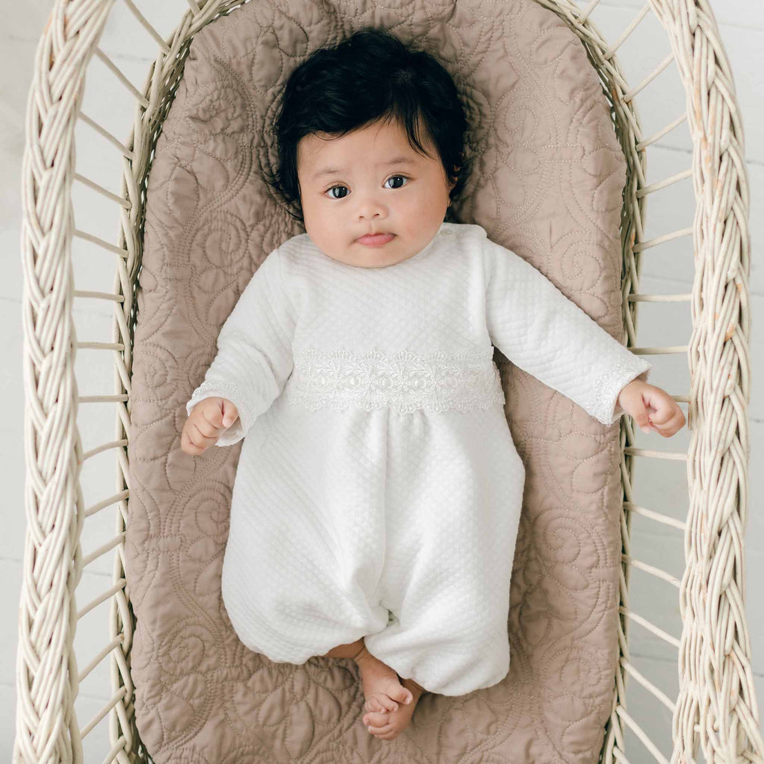 A baby with dark hair lies in a wicker bassinet, dressed in a Madeline Quilted Newborn Romper. The baby rests on a quilted cotton blanket, handmade in the USA, and looks up with a calm expression.