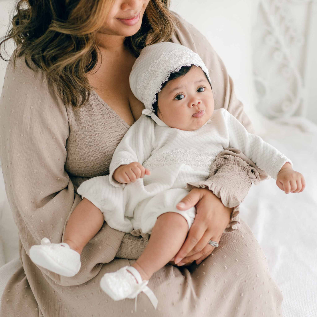 A woman gently cradles a baby dressed in a white Madeline Quilted Newborn Romper and bonnet. The baby, seated on her lap, looks attentively at the camera. The woman wears a beige dress with a subtle pattern and holds the baby's foot with one hand. Their expressions are calm and serene.