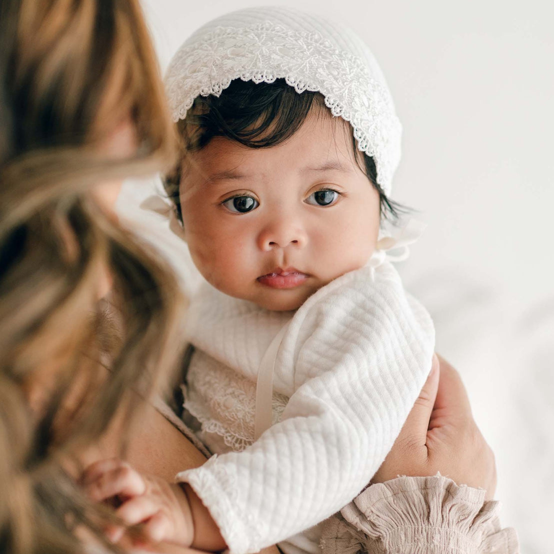 A baby wearing a white outfit and a delicate, handmade Madeline Quilted Newborn Bonnet with silk ribbon ties is being held by an adult. The baby's large, expressive eyes look directly at the camera. The adult's face is not visible, but they have wavy, shoulder-length hair. The background is softly blurred.