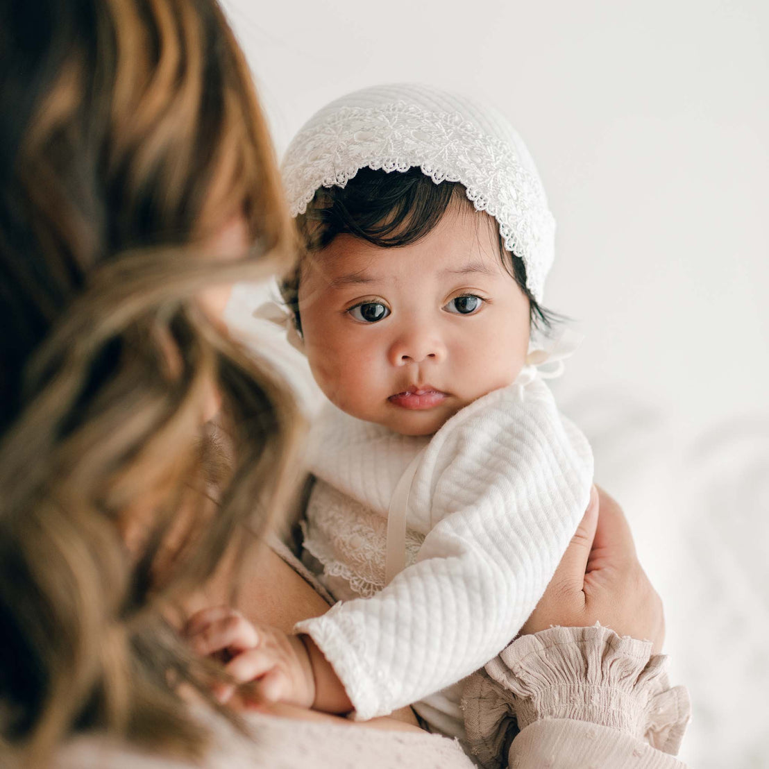 A close-up of a baby in a pristine, Madeline Newborn Gown and white lace bonnet, being held by an adult with wavy hair. The baby's big, dark eyes gaze directly at the camera, while the adult's face is turned away. The softly lit background adds a touch of warmth to the scene.