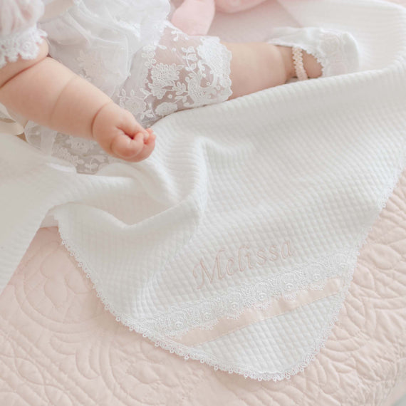 A baby in a delicate, handmade christening gown lies on a Melissa Personalized Blanket with intricate patterns. The personalized item features the name "Melissa" embroidered in light pink thread, accompanied by lace trim and a satin ribbon. The baby's hand and lace-trimmed socks are partially visible.