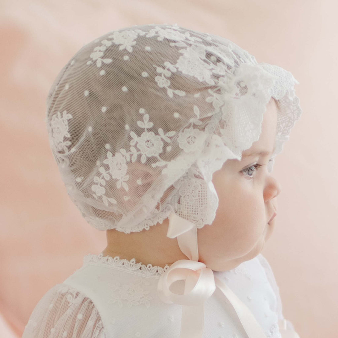 A baby is seen in profile wearing a Melissa Sheer Bonnet adorned with floral embroidered patterns and a ribbon tied under the chin. The infant also sports a matching white lace outfit. The background is a soft pink, providing a gentle, pastel ambiance.