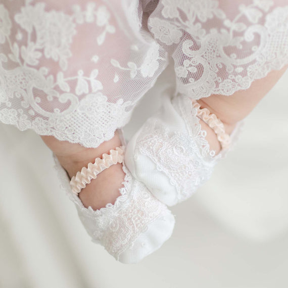 Close-up of a baby's feet dressed in delicate handmade Melissa Booties and white lace-trimmed clothing. The intricate floral patterns of the Venice lace create an elegant and soft appearance, with the 100% pima cotton ensuring comfort. The blurred background draws focus to the baby's feet.