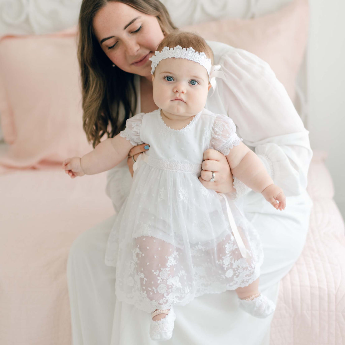 A woman in a white dress gently holds a baby dressed in a Melissa Romper Dress and headband. They are sitting on a pink bed, and the woman gazes lovingly at the baby, who looks forward with a curious expression.