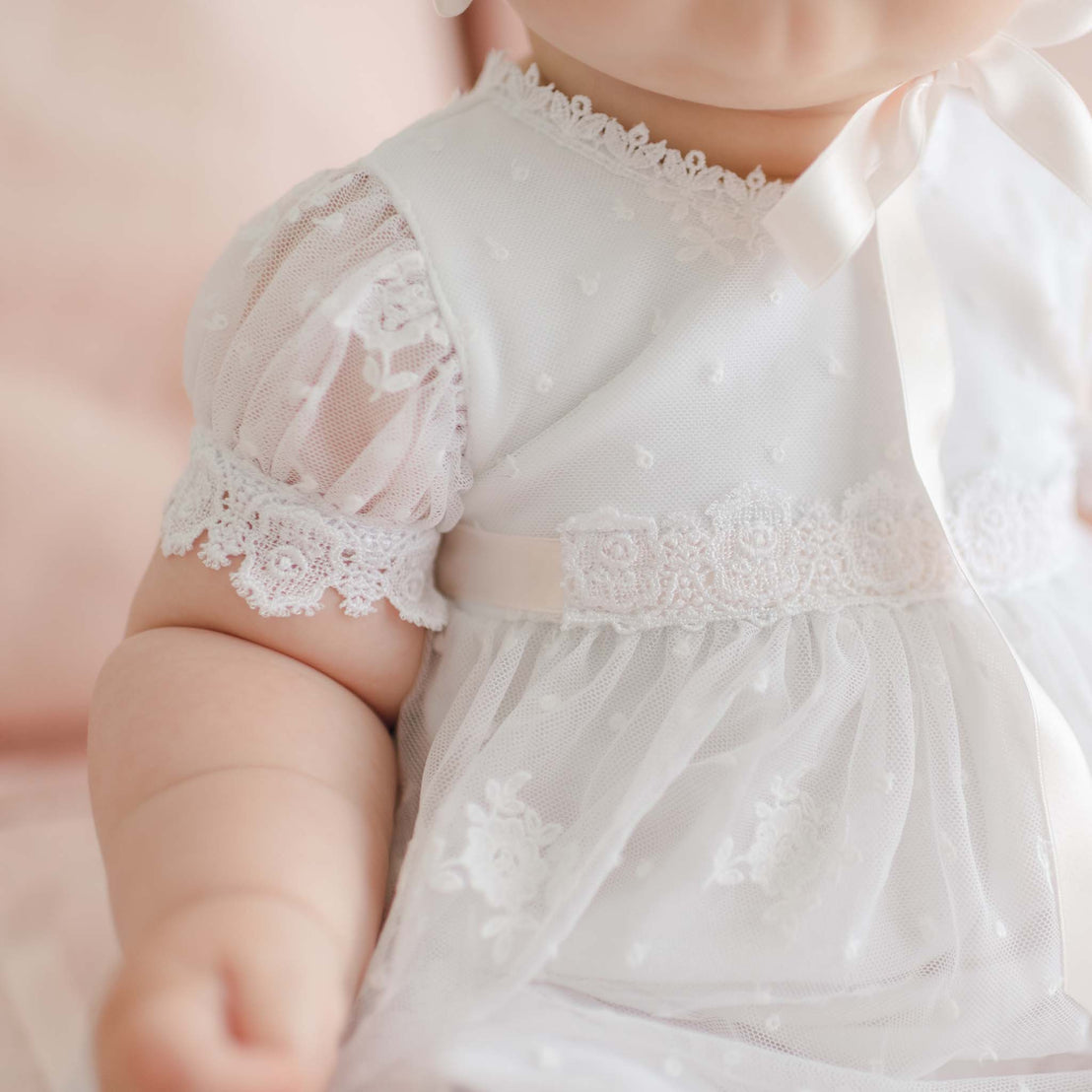 Close-up of a baby wearing a delicate white lace Melissa Romper Dress. The dress features sheer, puffed sleeves with intricate lace detailing and a ribbon bow at the neckline. The baby’s chubby arm is visible in the foreground, and the soft background hints at a light, cozy setting.