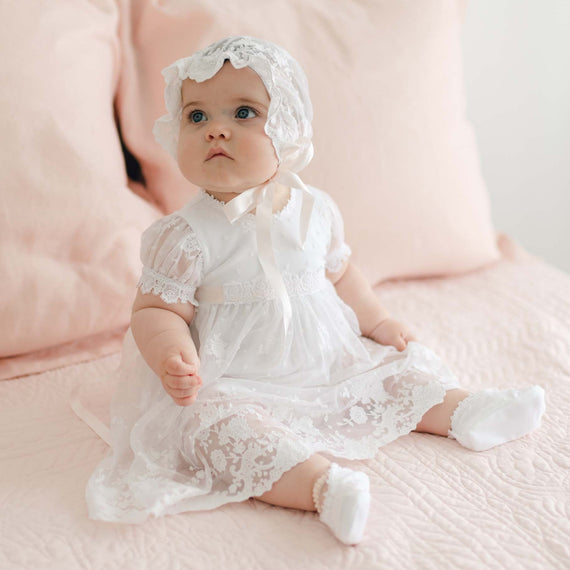 A baby wearing a delicate handmade white lace Melissa Romper Dress and matching bonnet sits on a pink quilted blanket with pillows in the background. The baby gazes upwards with a curious expression.