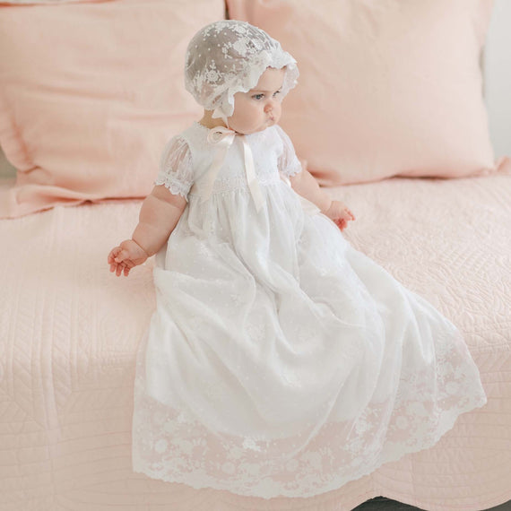 A baby wearing a long, white Melissa Christening Gown & Bonnet adorned with delicate Venice lace trim sits on a light pink quilted bedspread. The baby looks off to the side, and the background includes blush pink pillows.