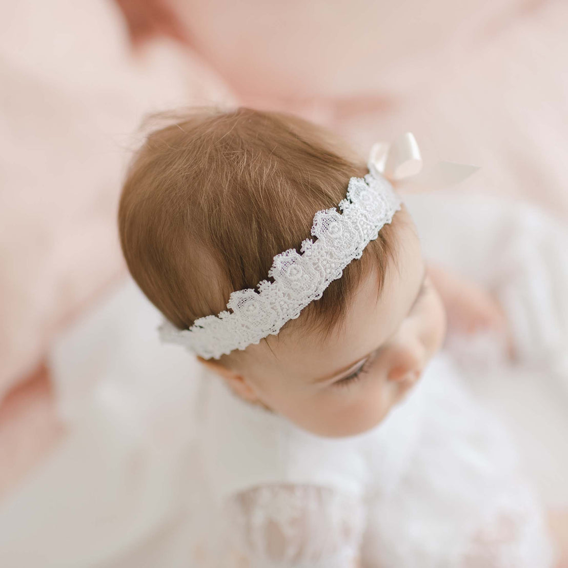 A baby with light brown hair wears a delicate Melissa Headband with a small silk ribbon bow, dressed in a pristine white outfit. The photo is taken from above and slightly to the side, set against a soft pink and white background, creating a serene and gentle ambiance.