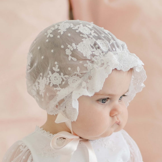 A baby dressed in white is wearing a delicate, lacy Melissa Sheer Bonnet adorned with a floral embroidered pattern and ruffles. The baby has a serious expression and is seen against a blurred, soft pink background.