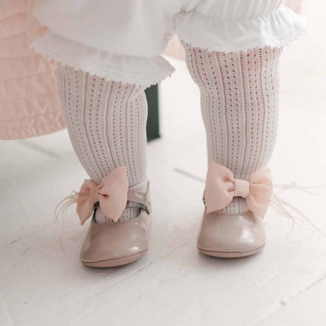 A close-up of a toddler's legs showcases the Openwork Fancy Tights with their delicate, intricate design and light pink patent leather shoes featuring charming pink bows. The child also wears white ruffled bloomers for an added dressy touch while standing on a white floor.
