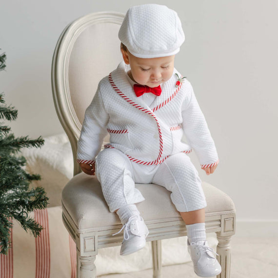 A toddler in an adorable Nicholas 3-Piece Suit & Bow Tie Set, showcasing a white ensemble with red accents and a matching bow tie, sits on a chair next to the Christmas tree. Wearing a cap and quilted cotton suit handmade in the USA, the child gazes down at their feet amidst wrapped gifts and cozy ambience.