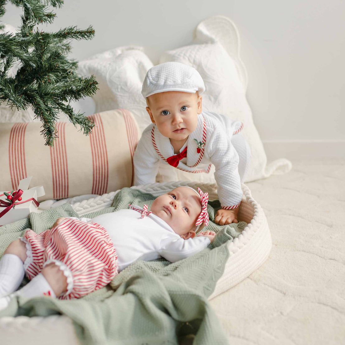 Two babies dressed in the Nicole Bloomer Set are near a decorated Christmas tree. One baby is lying on a pillow basket, while the other, in a red and white striped cotton onesie set and cap, sits nearby. The setting is cozy with a striped cushion and soft blanket.