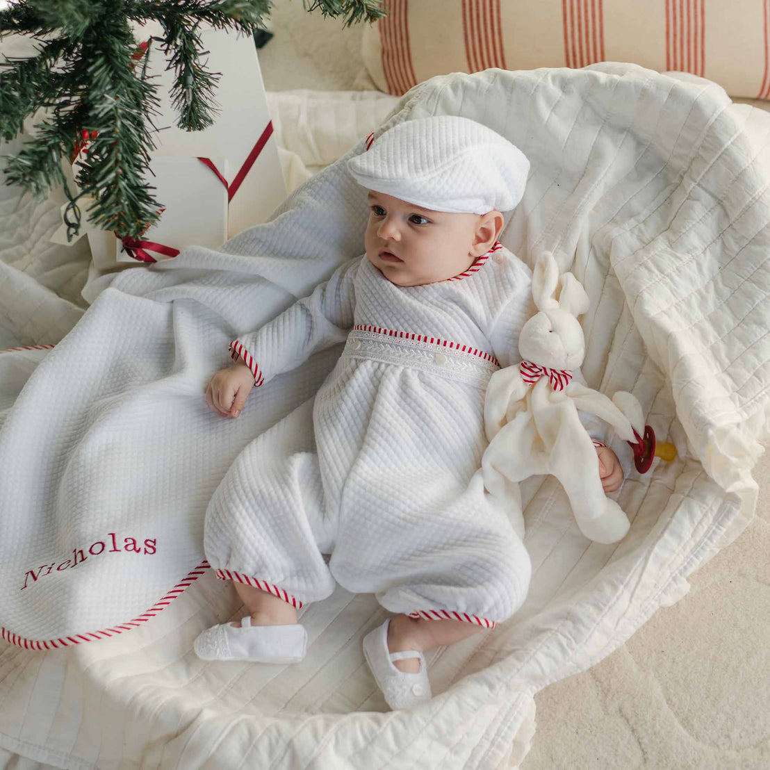 A baby dressed in a handmade Nicky Quilted Romper with red accents lies on a cozy, cream-colored blanket. The baby holds a plush white rabbit and is next to a personalized blanket with "Nicholas" embroidered in red. A tree branch is visible above, adding to the serene holiday season vibe.