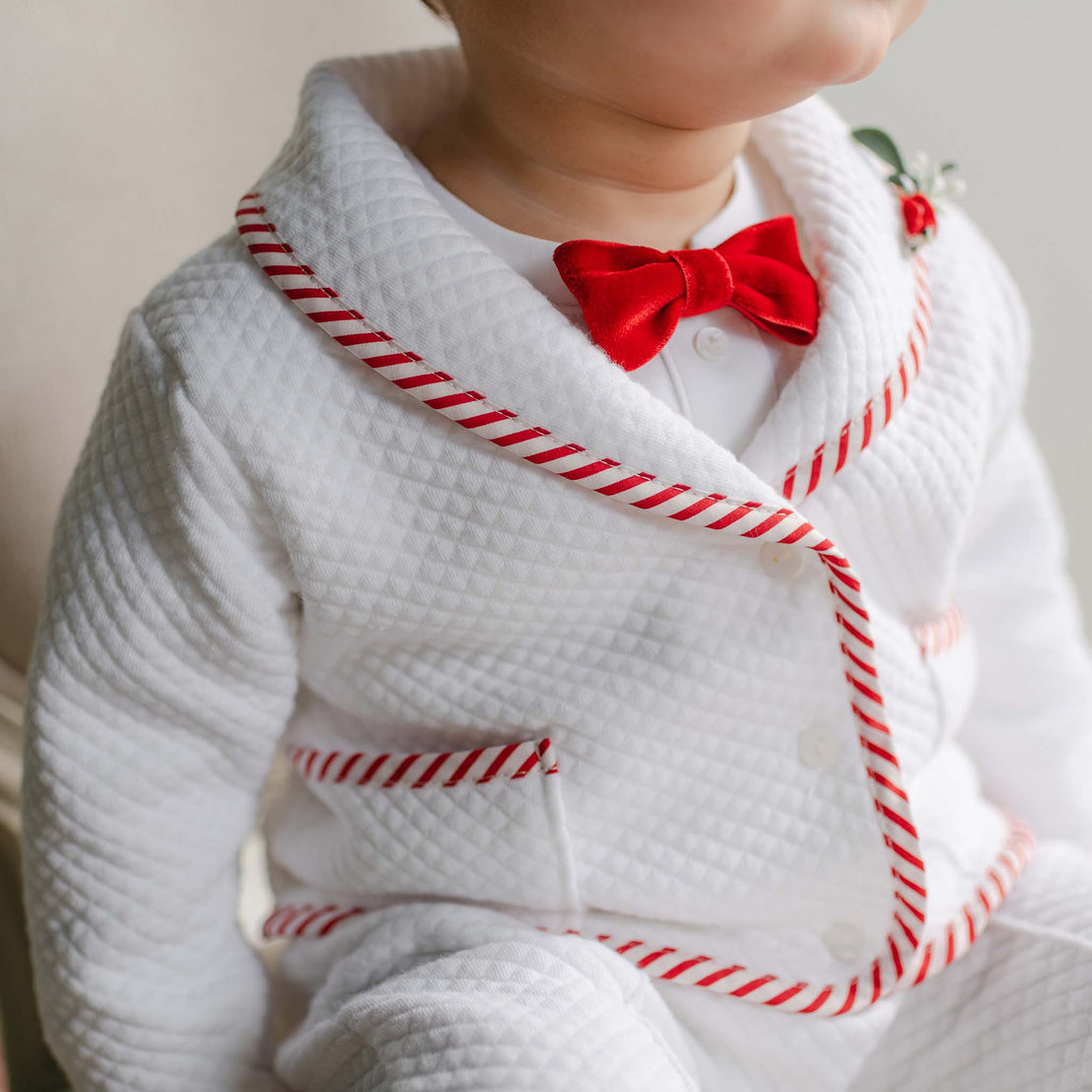 A young child is elegantly dressed in the Nicholas 3-Piece Suit, featuring a quilted cotton design with red trim and a charming red bow tie, making it perfect for Christmas. This suit, which could be handmade, includes red accents on the collar and pockets and is adorned with a small decorative plant pin on the lapel. The child sits comfortably indoors.