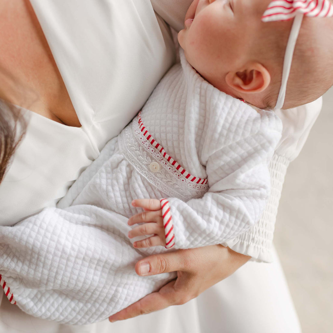 A baby dressed in the Nicky Quilted Romper with red-striped accents and a matching headband is cradled in an adult's arms. The adult, dressed in white, gently holds the child close.