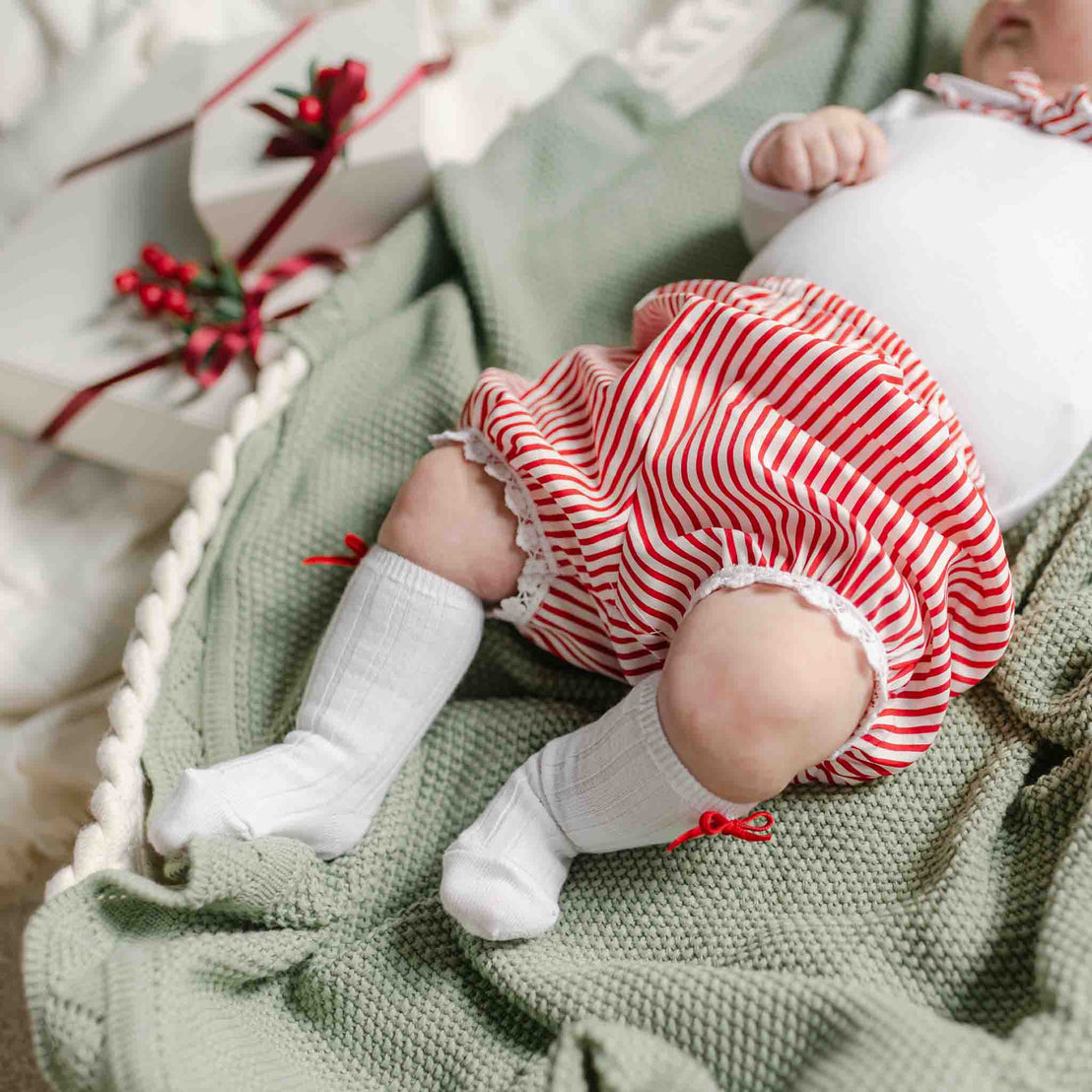 A baby dressed in the Nicole Bloomer Set, featuring cheerful red and white striped bloomers, white long socks adorned with red bows, and a crisp white top is lying on a green knitted blanket. Nearby, there's a charming white gift box with a vibrant red ribbon, making it ideal for holiday occasions. A coordinating bow headband perfectly completes this adorable outfit.