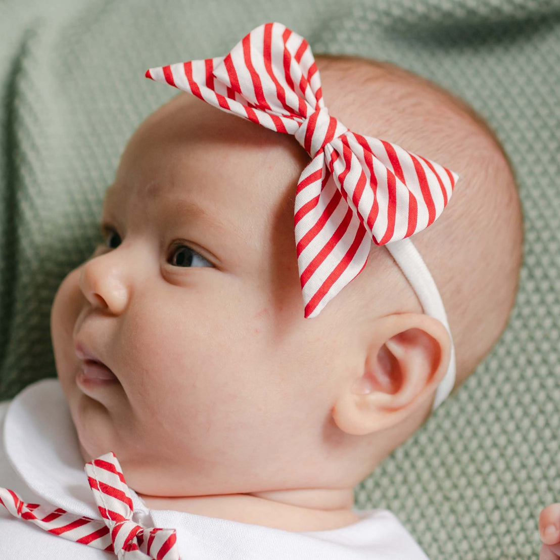 A baby wearing a red and white plaid bow hair clip peacefully rests on a green textured blanket, dressed in a white outfit featuring a small, handmade bow crafted in the USA.