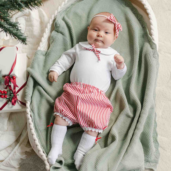 A baby lying in a basket, dressed in the Nicole Bloomer Set featuring red and white striped bloomers with a matching bow headband, ready for holiday events.