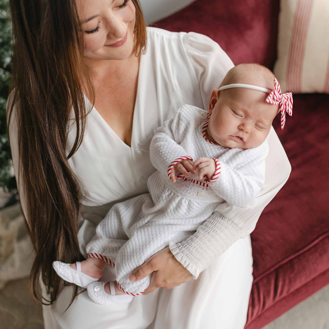 A woman with long hair, attired in a white ensemble, gently holds a sleeping baby on a red sofa. The baby is peacefully resting in her arms while wearing a striped cotton outfit featuring red details and the Nicole Bow Headband crafted from nylon.