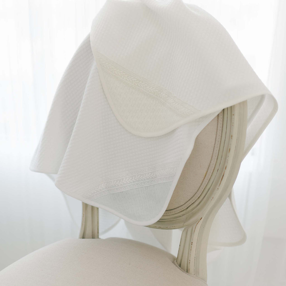 Two Oliver Personalized Blankets, one in white and the other in ivory, are draped over the back of a cream-colored chair with a curved backrest. The Oliver Personalized Blanket features intricate, subtle patterns near the edges and the corner of the blanket. The background is softly lit.