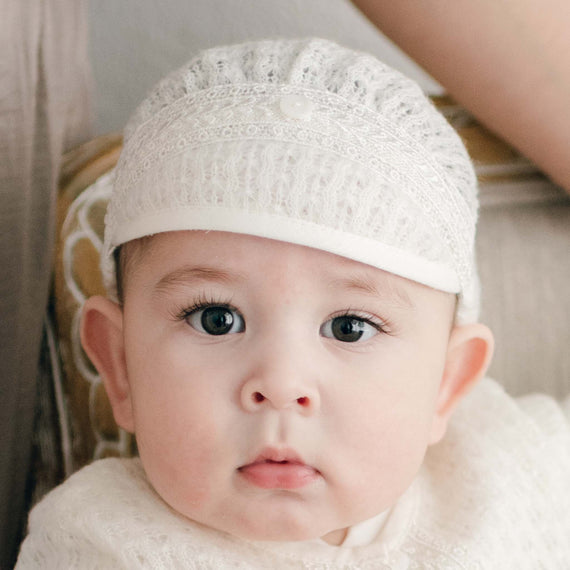 A baby boy wearing the Oliver Knit Cap in ivory. The cap is made from a luxurious material with a beautiful texture. It features a delicate linen and cotton border and Venetian lace with a button in the center. The boy looks ahead with a curious expression.