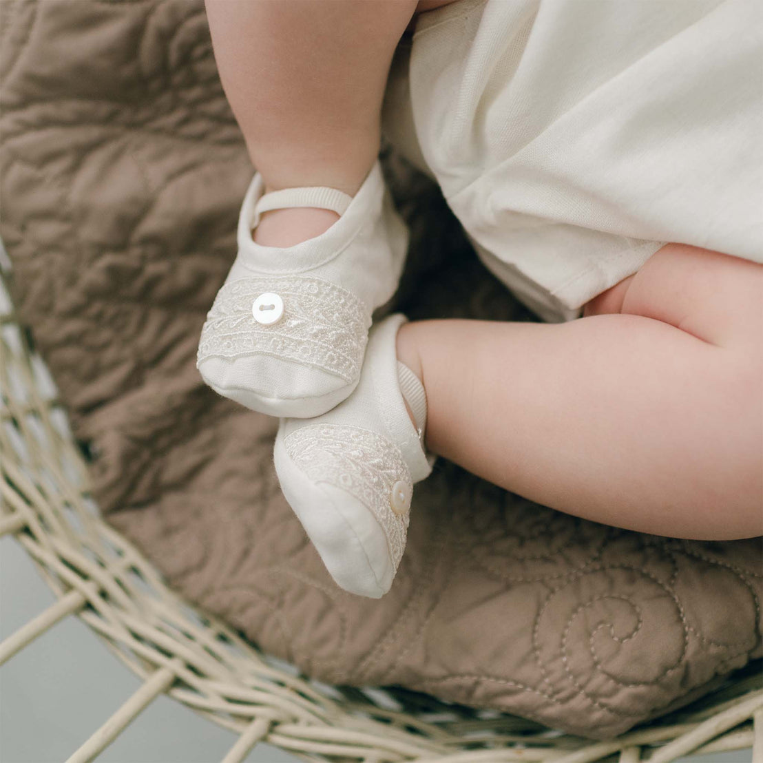 The top detail of the Oliver Booties in ivory worn by a baby boy. The Oliver booties are made with a linen top, soft pima cotton bottom, and features a Venice lace and button detail (with elastic strap across the foot). The background is a wicker basket lined with a brown quilted blanket with fleur-de-lys design.