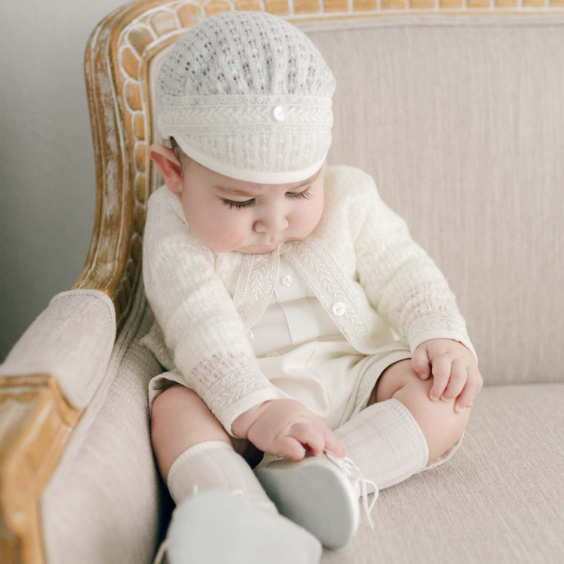 A baby dressed in the Oliver Romper and Knit Sweater and Cap ensemble in ivory featuring intricate embroidery. He is sitting on a light-colored upholstered armchair. The baby is looking down and seems to be touching their white shoe.