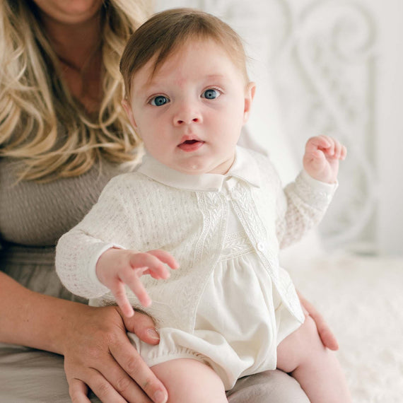 A baby boy with fair skin and light brown hair, dressed in an Oliver Knit Sweater, sits on a woman's lap. The woman with long blonde hair is wearing a light gray dress. They are indoors with a softly lit background. The baby looks directly at the camera with a curious expression.